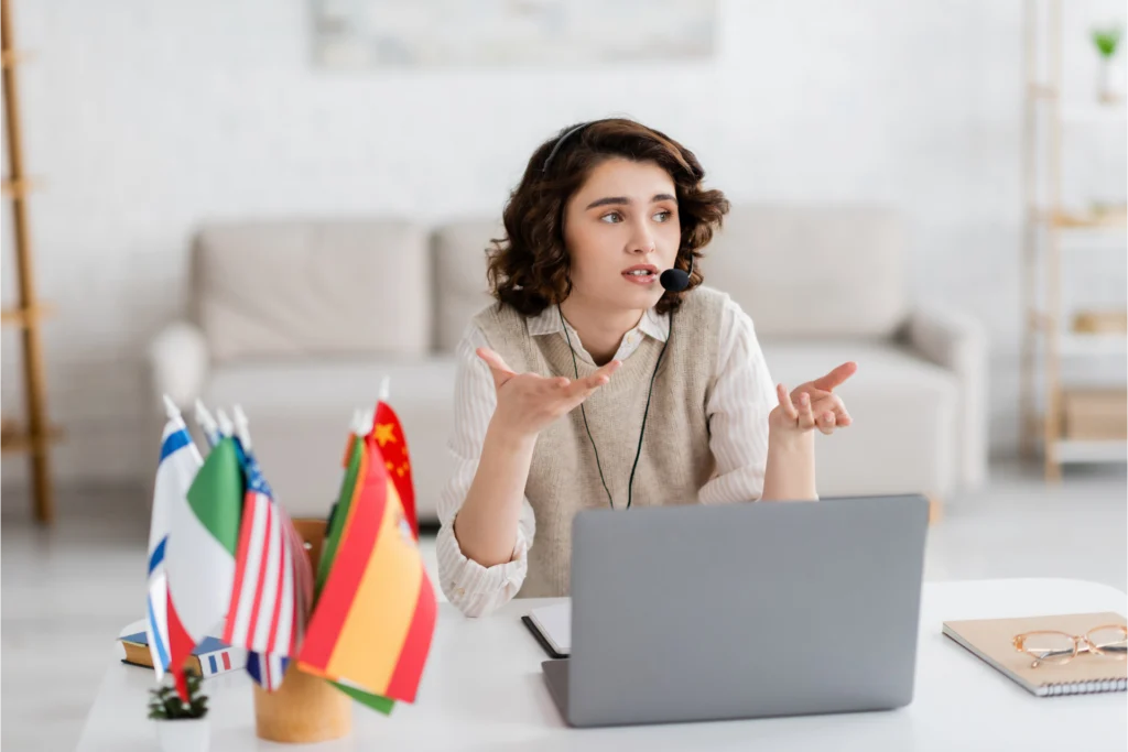 brunette-language-teacher-headset-gesturing-near-laptop-blurred-international-flags-home