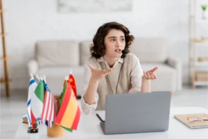 brunette-language-teacher-headset-gesturing-near-laptop-blurred-international-flags-home